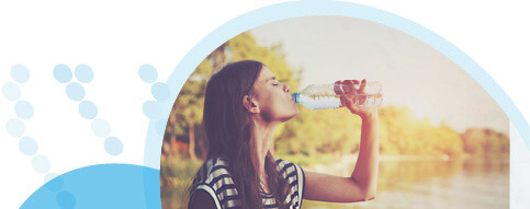 אישה עם שיער חום וחולצת פסים שותה מים מבקבוק על רקע מקווה מים ועצים